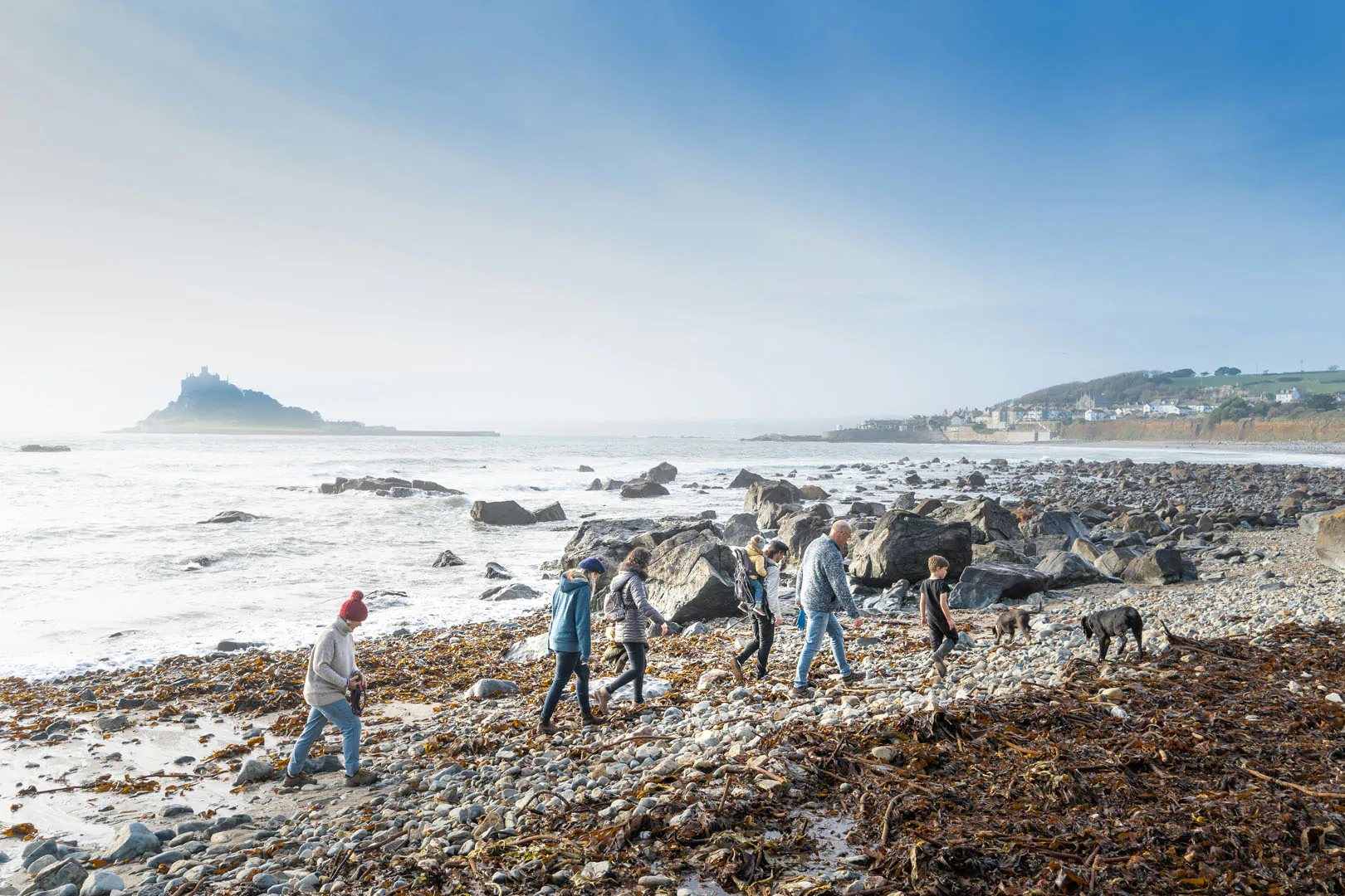 People walk on the beach with a dog, taking in the beautiful surroundings of St Michael's Mount in Cornwall.