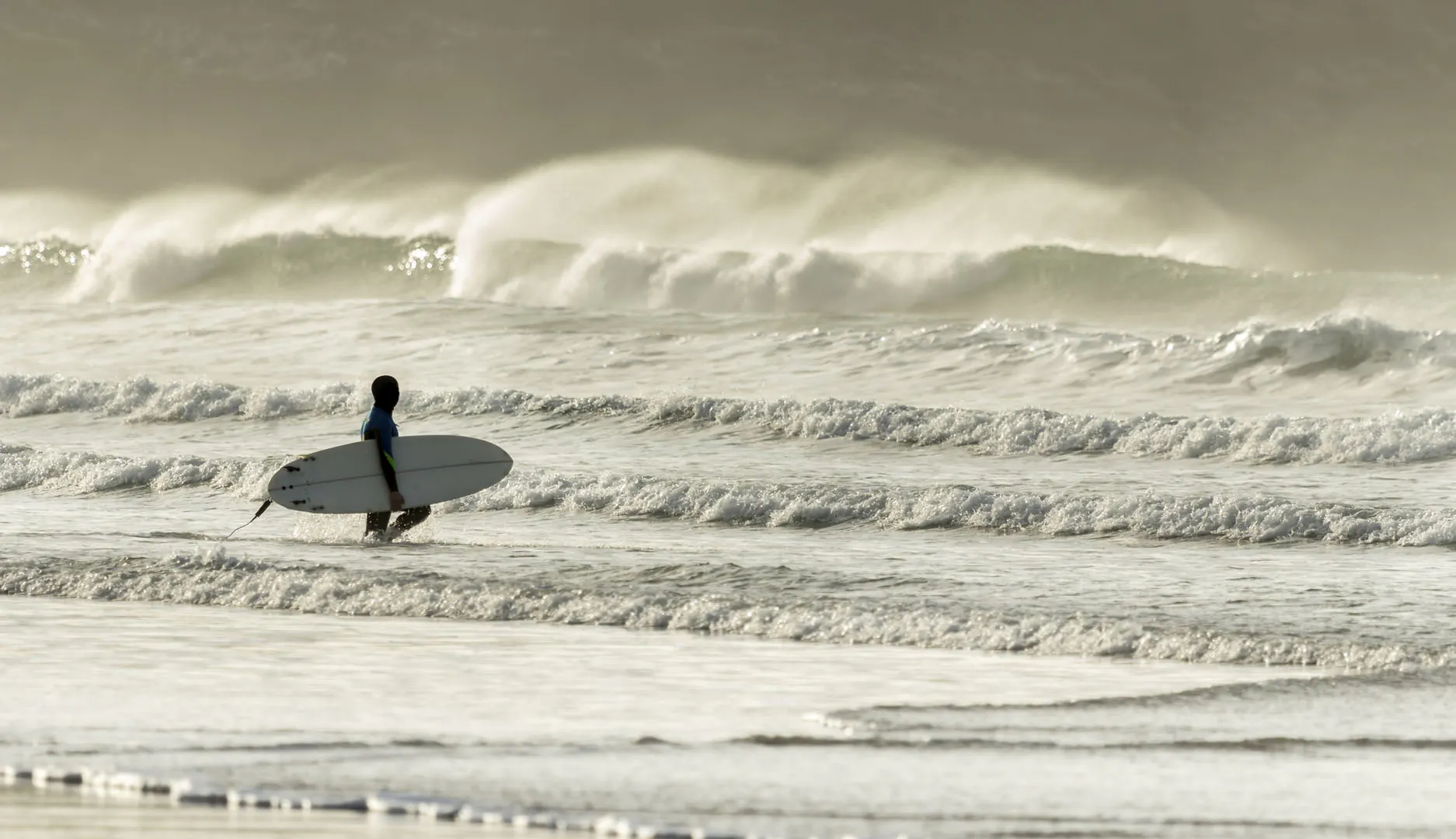 A person walks along Fistral Beach, carrying a surfboard, ready to catch waves at Cornwall's best surf spot.