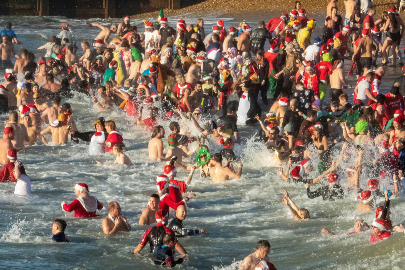 A festive crowd in Santa hats enjoying a Christmas sea swim in Cornwall, splashing in the water under a clear sky.
