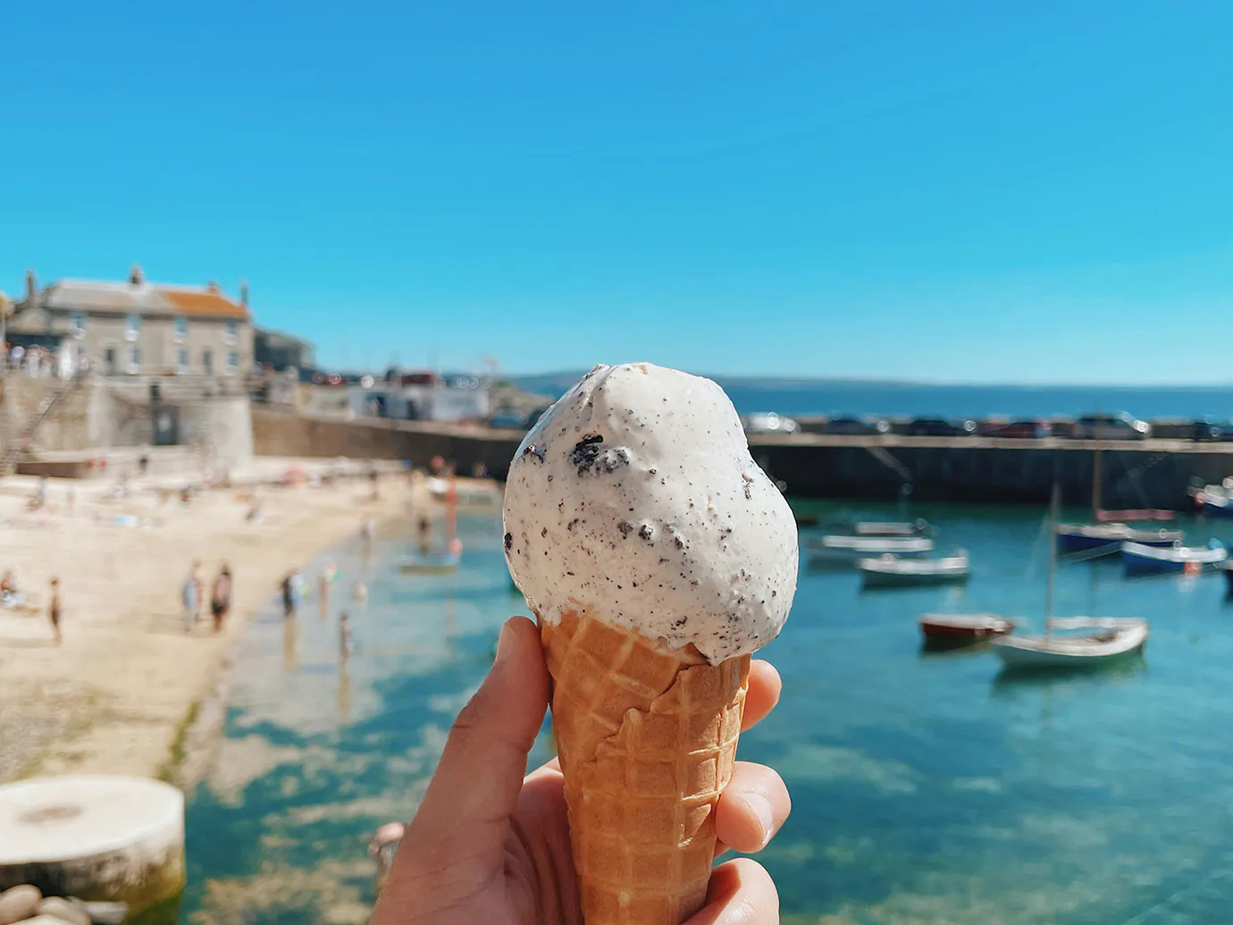 A person enjoys a colourful local ice cream cone by the water in Mousehole, Cornwall, with a scenic view in the background.